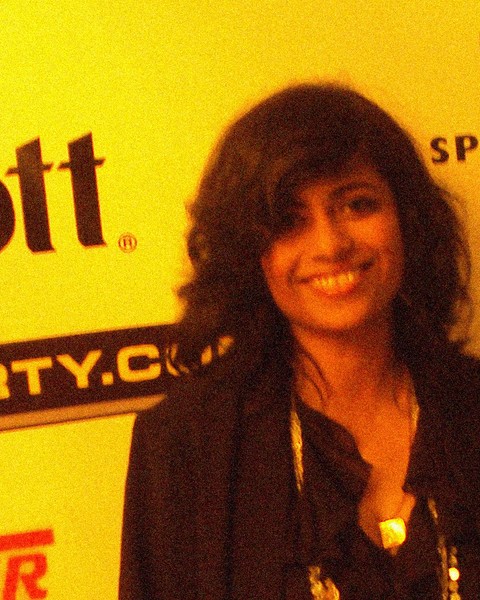 Grainy photo: fine speckles and colored dots on a smiling face, dark hair, and a yellow backdrop